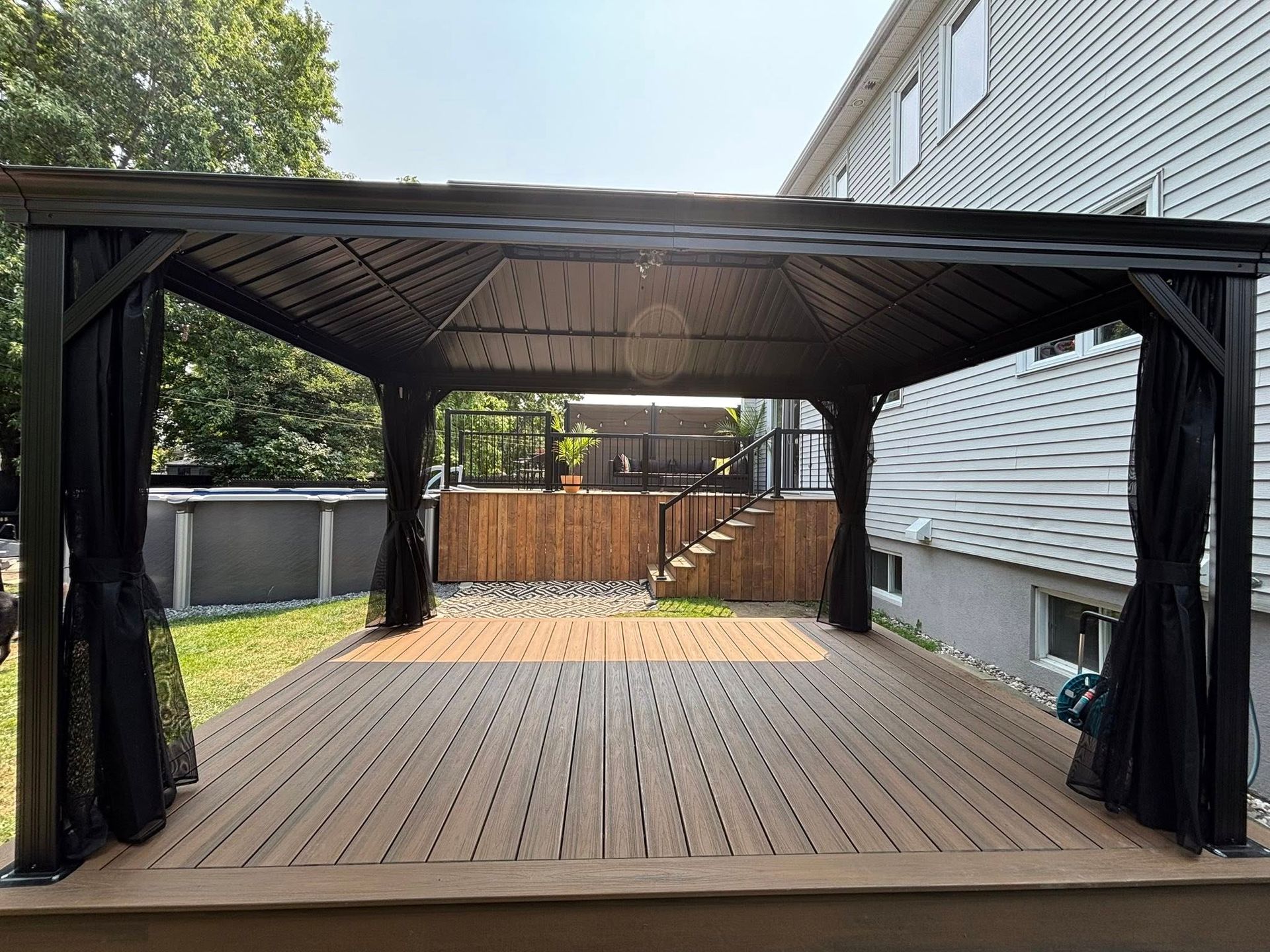 Gazebo avec rideaux sur une terrasse en bois donnant sur un jardin avec piscine et escaliers menant à un niveau supérieur.