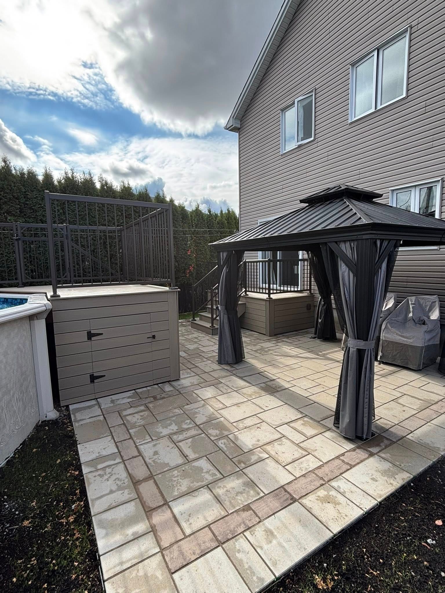 Terrasse avec gazebo et dalles de pierre à côté d'une maison et d'une piscine, sous un ciel nuageux.