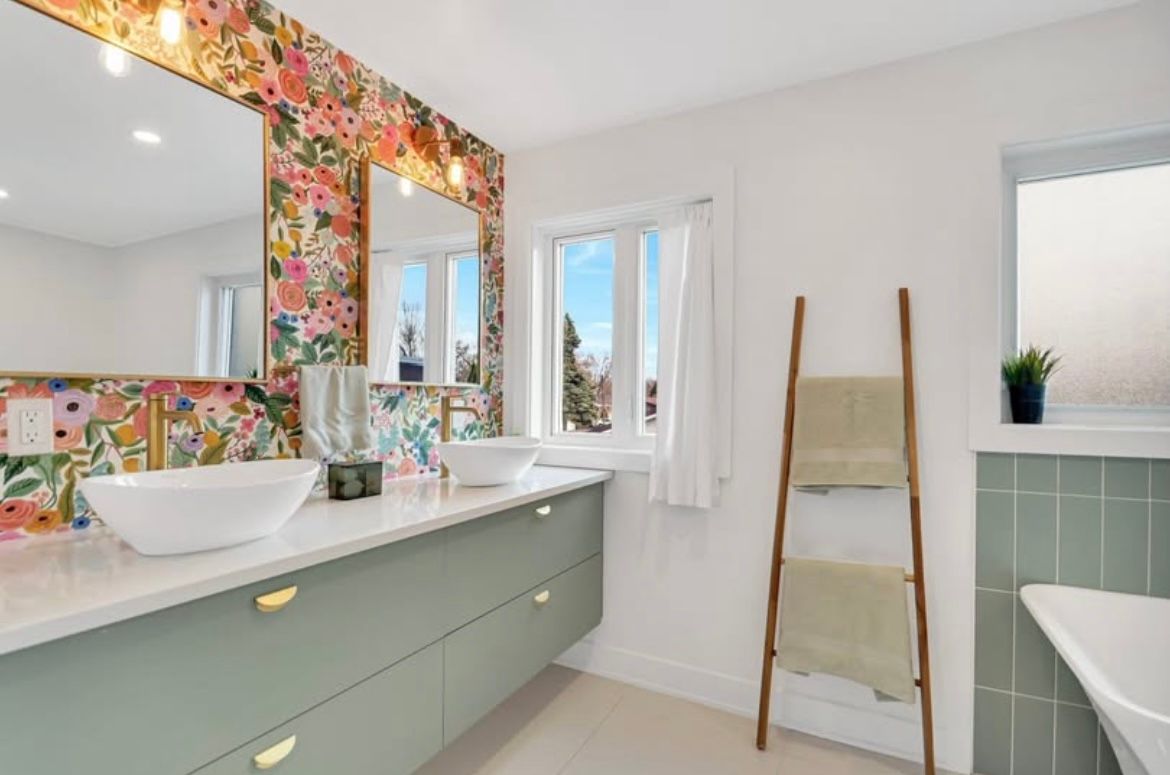 Salle de bains avec papier peint à motifs floraux, meuble-lavabo vert, lavabos blancs et échelle porte-serviettes en bois.