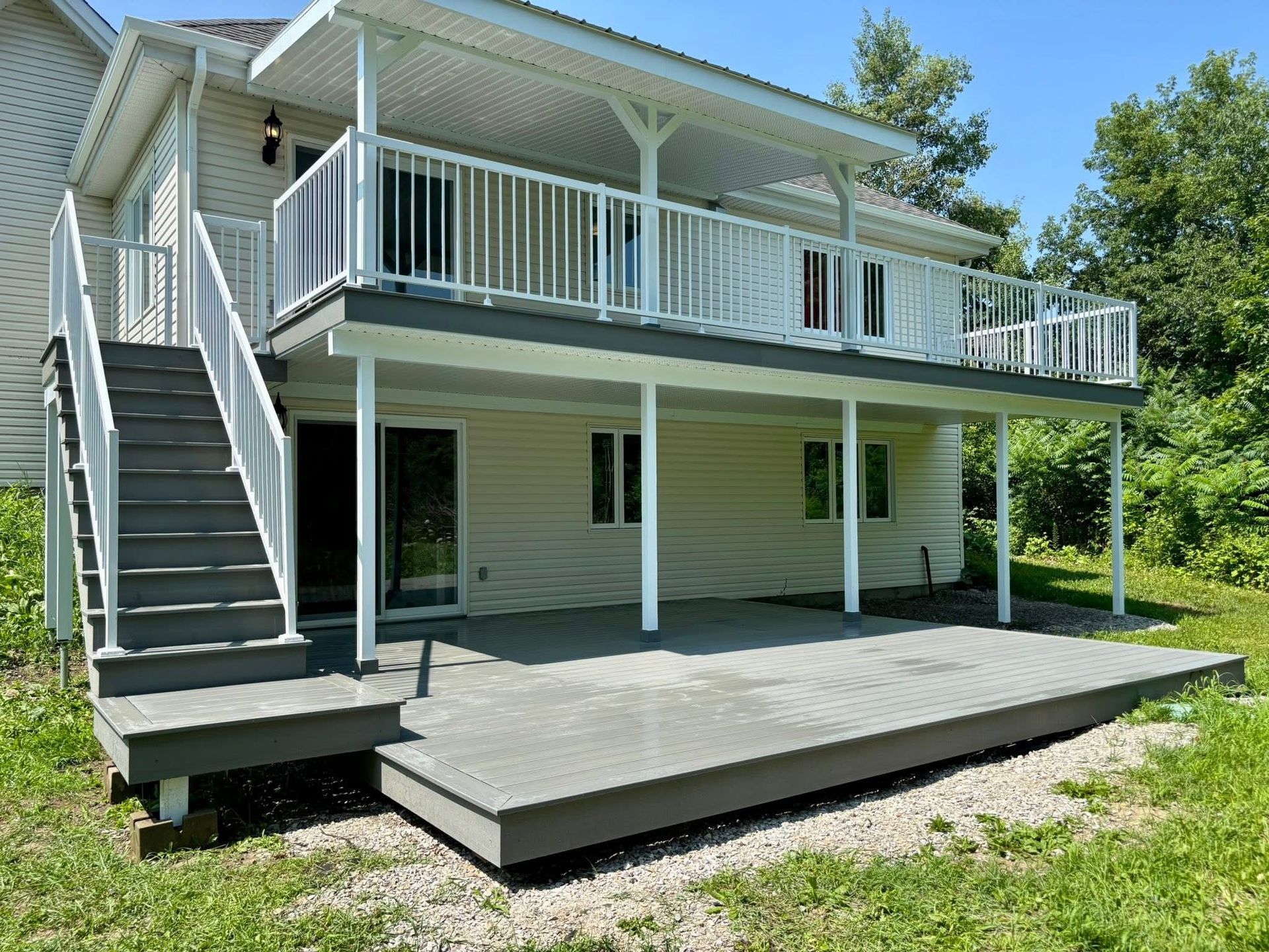 Maison à deux étages avec terrasse et escalier gris. La terrasse est soutenue par des piliers blancs. Journée ensoleillée.