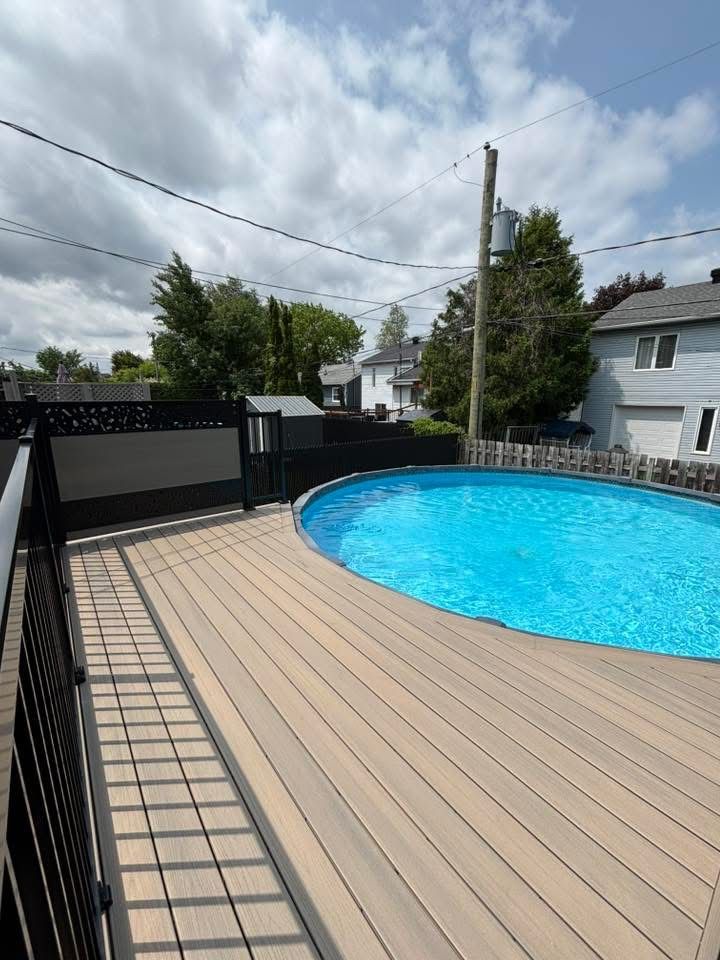 Piscine hors sol avec terrasse en bois, clôture noire et poteau électrique dans un jardin sous un ciel nuageux.