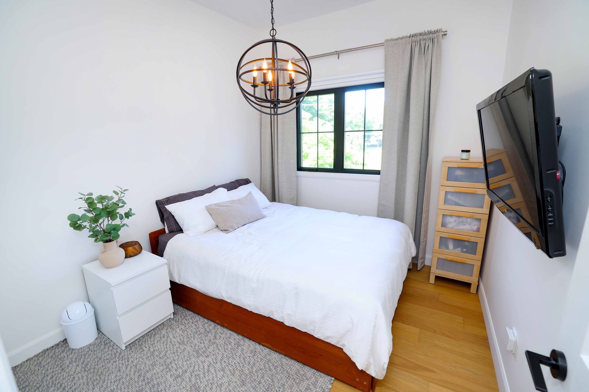 Chambre avec murs blancs, lit, fenêtre, télévision et luminaire décoratif.