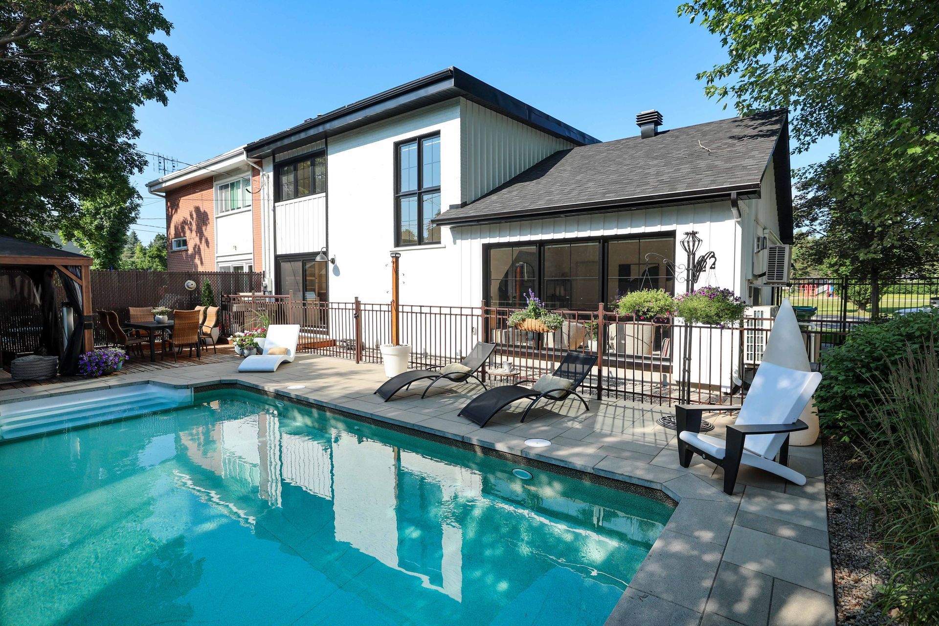Jardin avec piscine, maison blanche moderne, chaises longues et clôture. Ciel bleu.