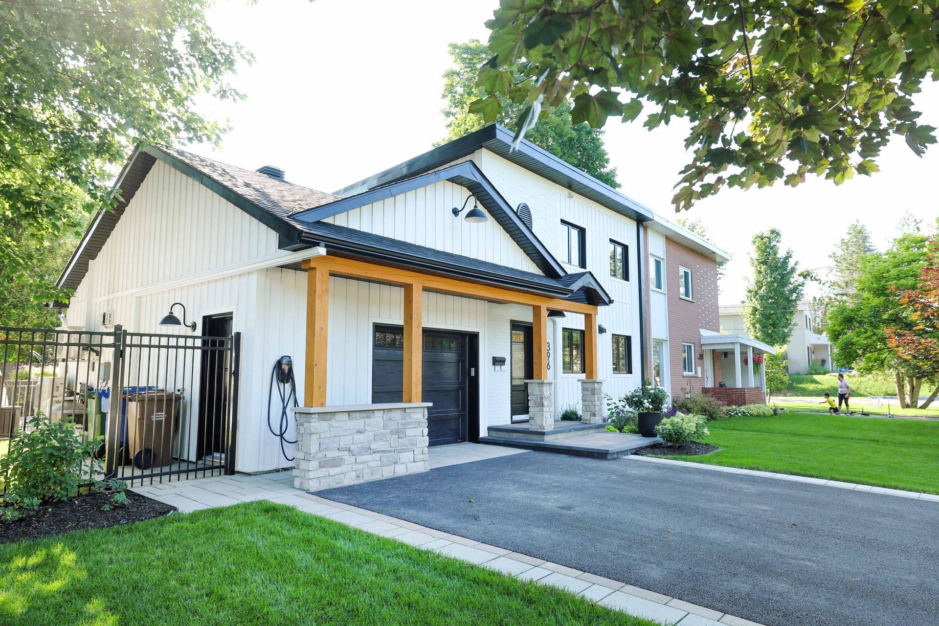 Maison à deux étages, avec allée privée. Porche avec poutres apparentes, pelouse verte, arbres. Journée ensoleillée.