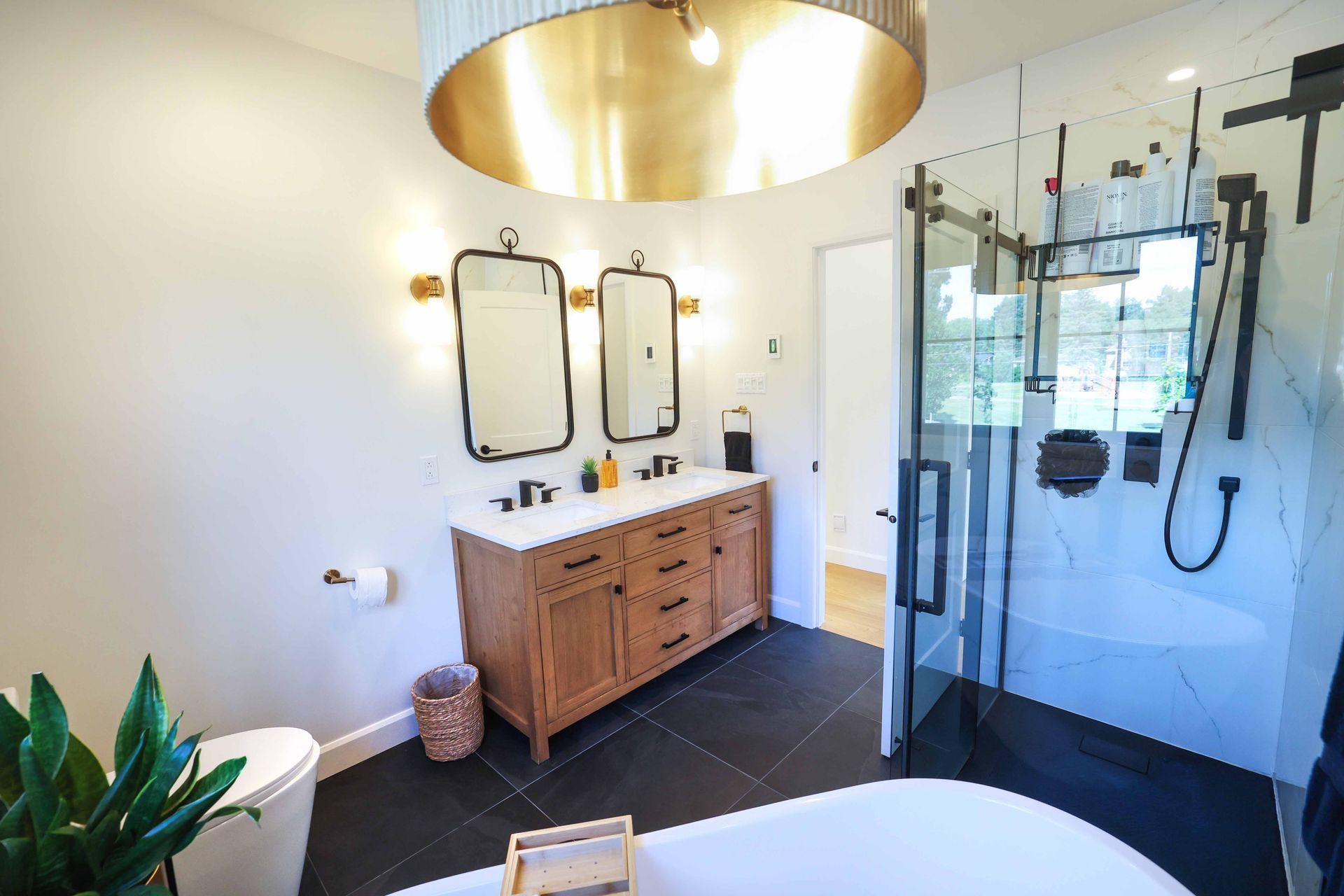 Salle de bains avec meuble-lavabo en bois, sol en carrelage noir, douche en verre et luminaire doré.