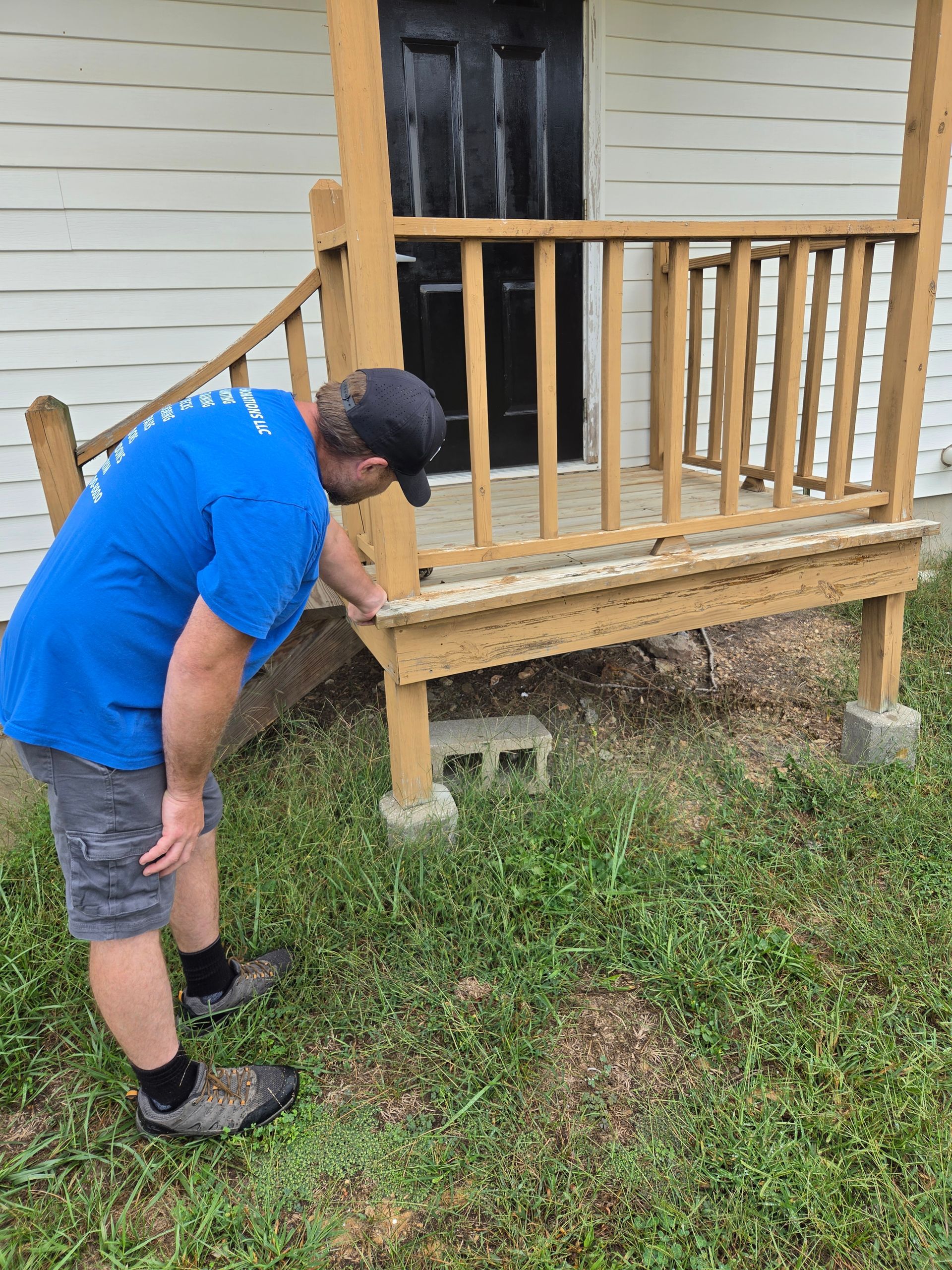 Man inspecting the underside of a wooden porch with railings beside a house.