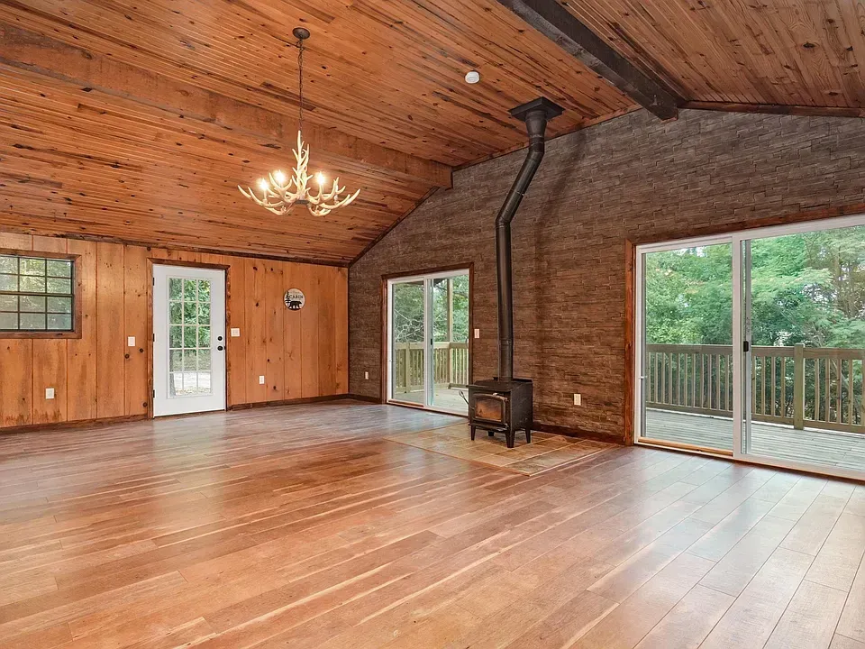 Spacious wood-paneled living room with vaulted ceiling, chandelier, wood stove, and sliding glass doors to a deck