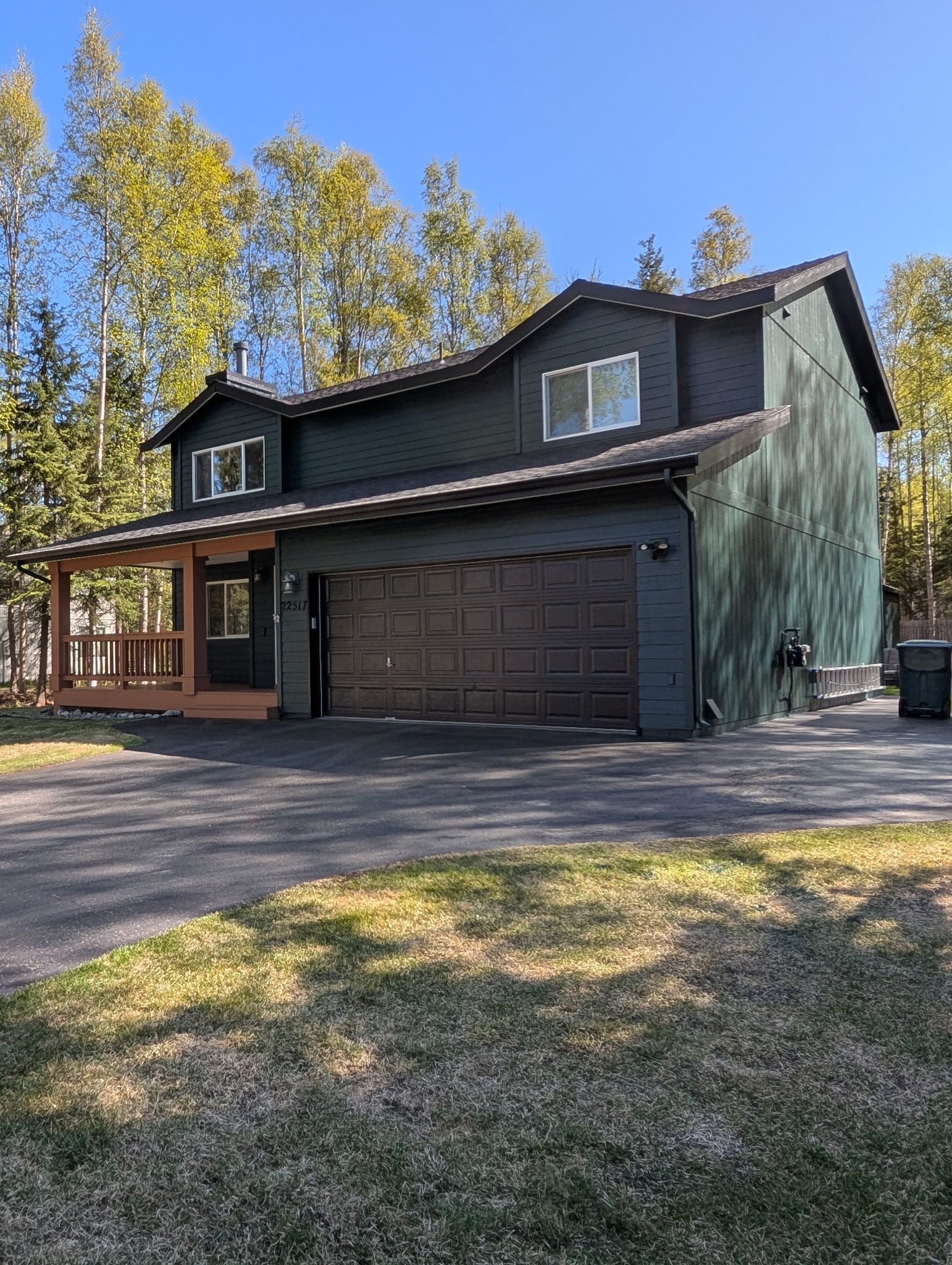 Dark two-story house with a double garage and front porch, set among tall trees on a sunny day