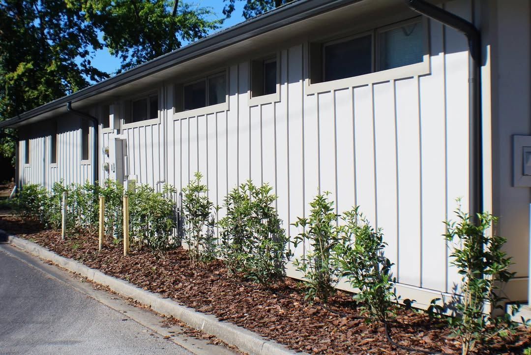 Row of light-colored buildings with vertical siding, small green bushes planted in a bed of brown mulch.