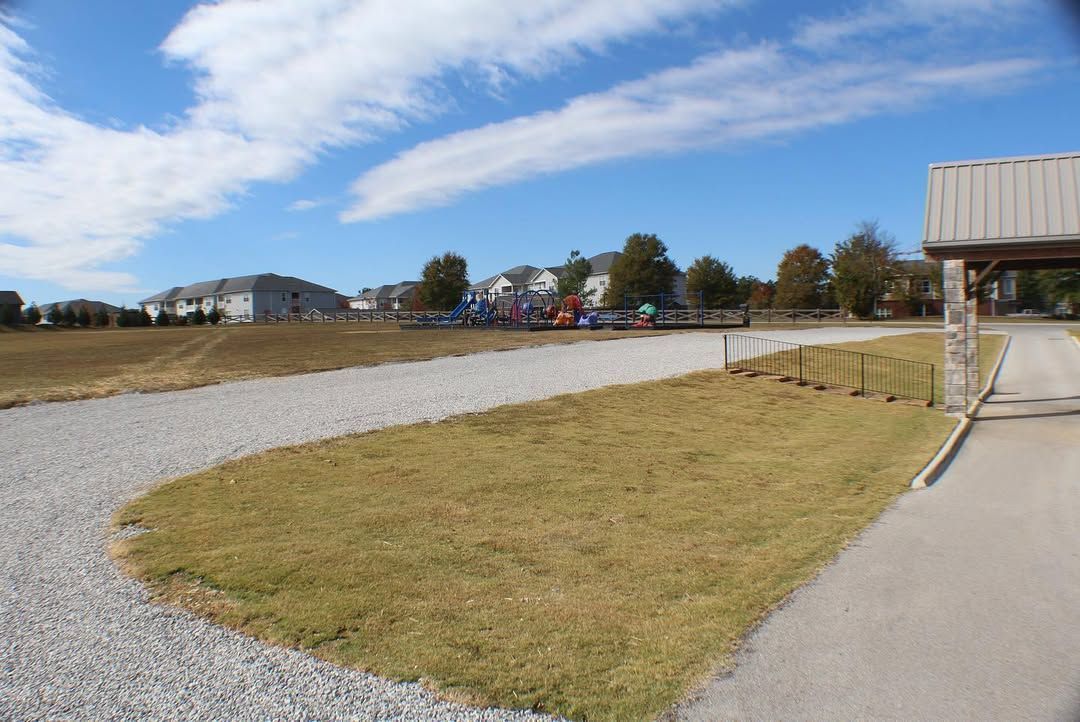 Gravel path curves past a grassy area, leading to a playground and buildings on a sunny day.