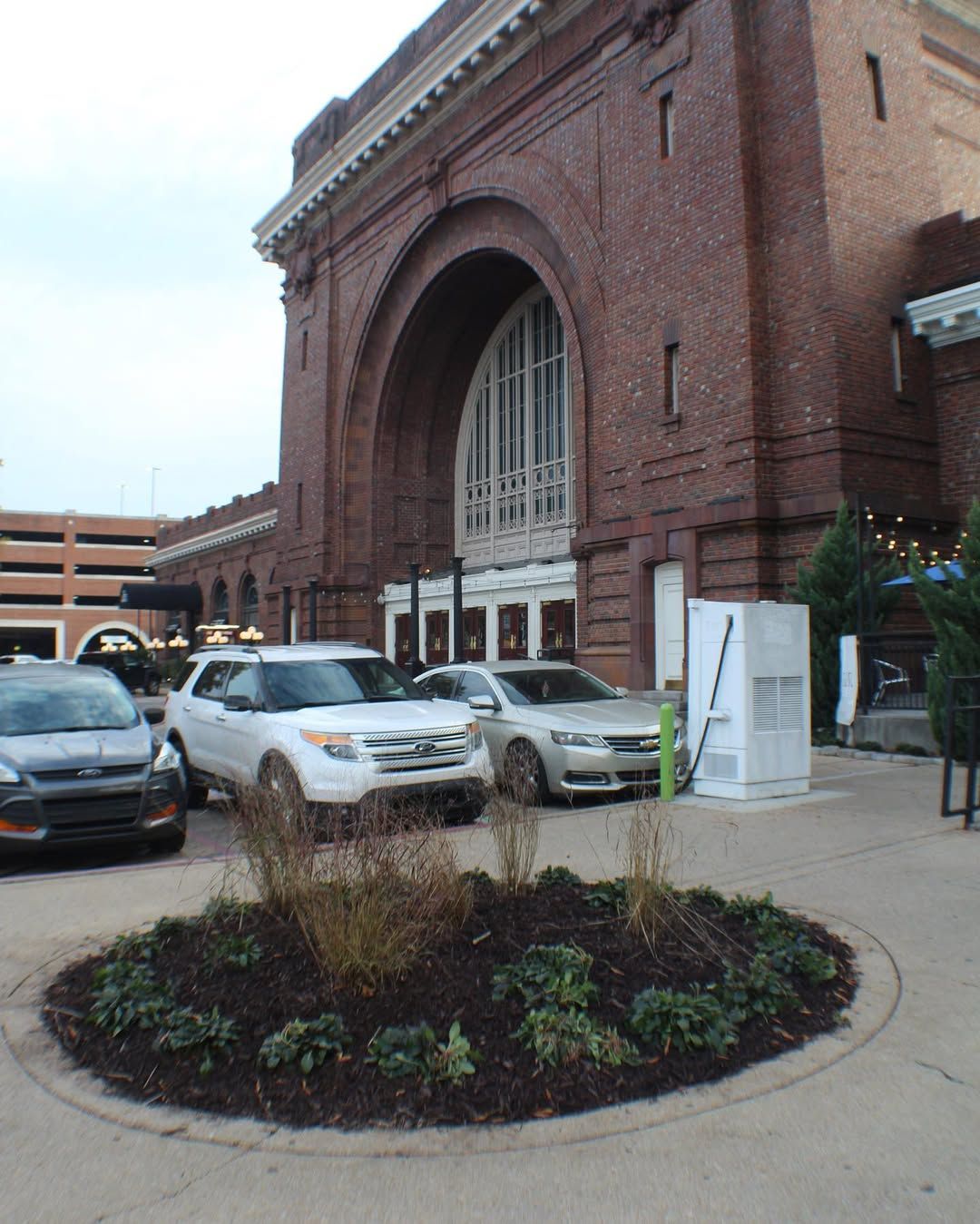 Exterior view of a brick building with arched entrance, cars parked in front, and a circular garden bed.