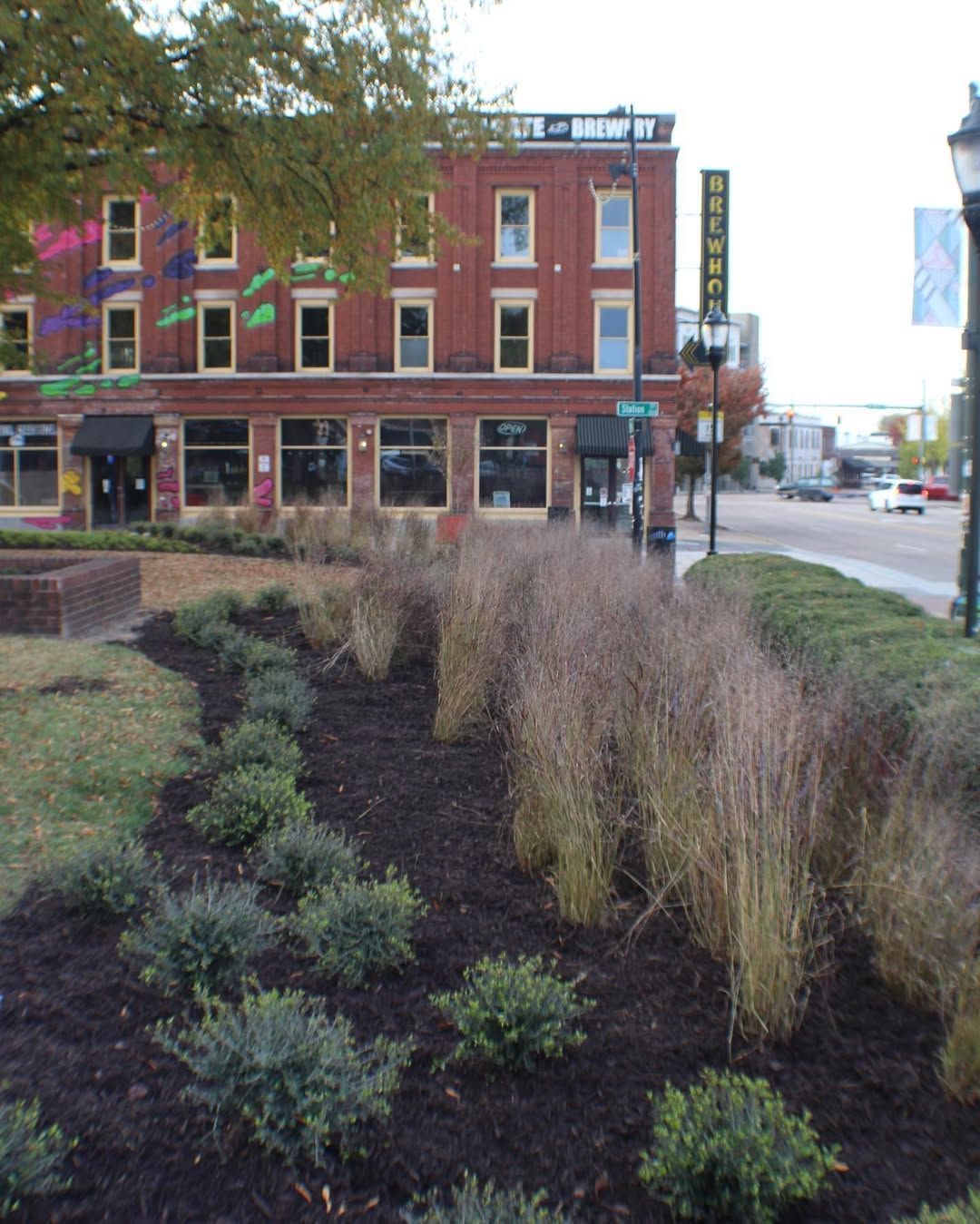 Brick building with ground-level storefronts, brown mulch beds with plants, and a street view with a car.