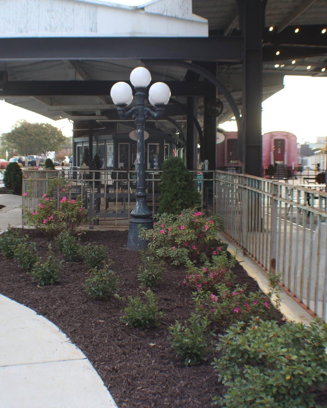 A black lamppost with 3 globes stands in a landscaped area near a covered seating area, with a train car in the background.