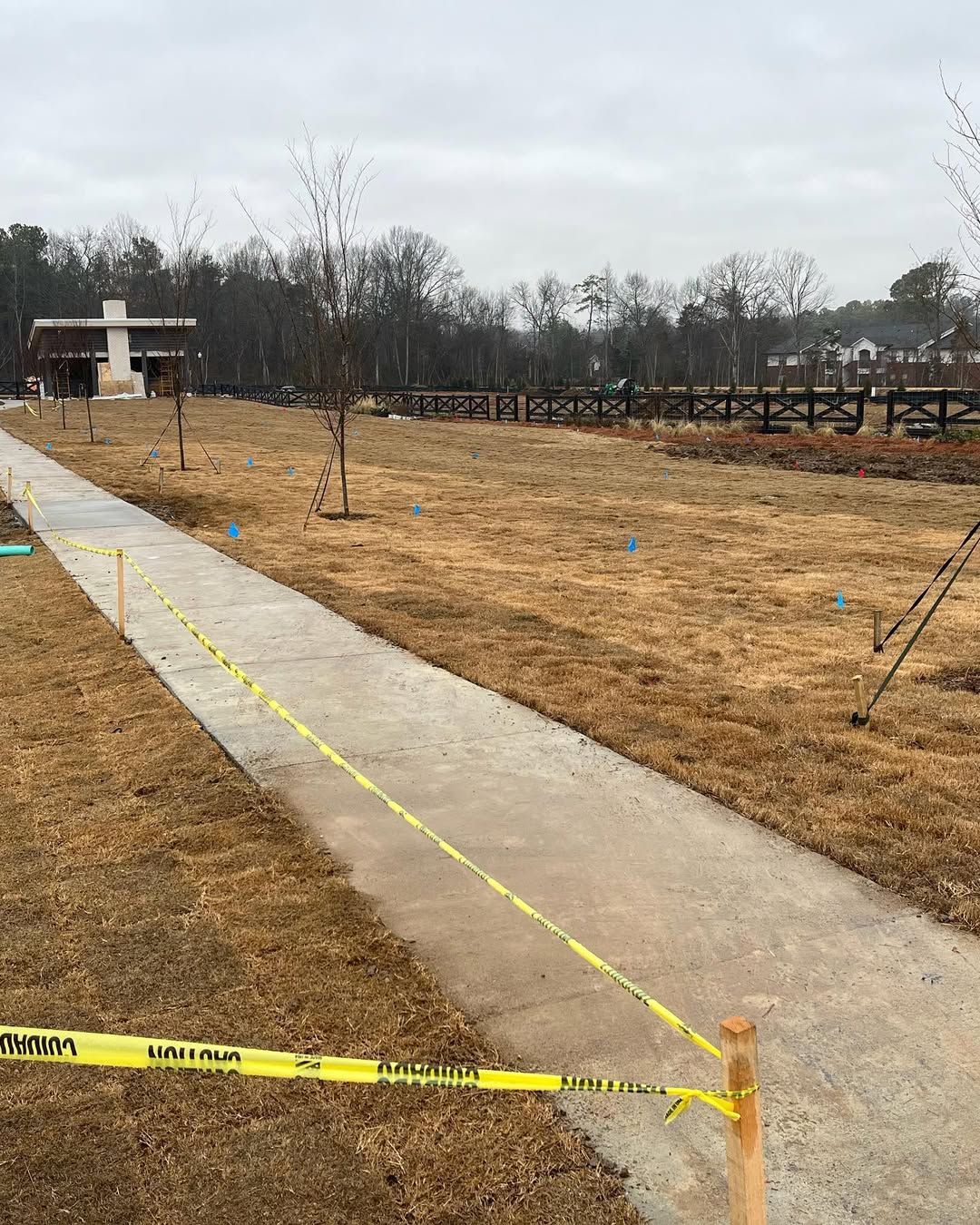 Concrete path in park with yellow caution tape, brown grass, and trees. A building and fence are visible.