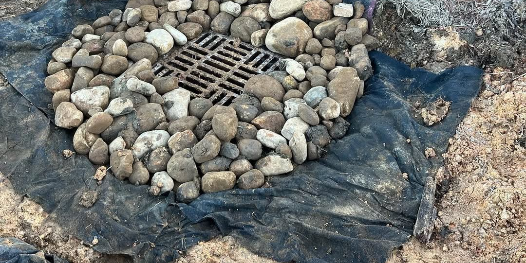 Rock-lined drain grate set in black plastic sheeting on bare ground
