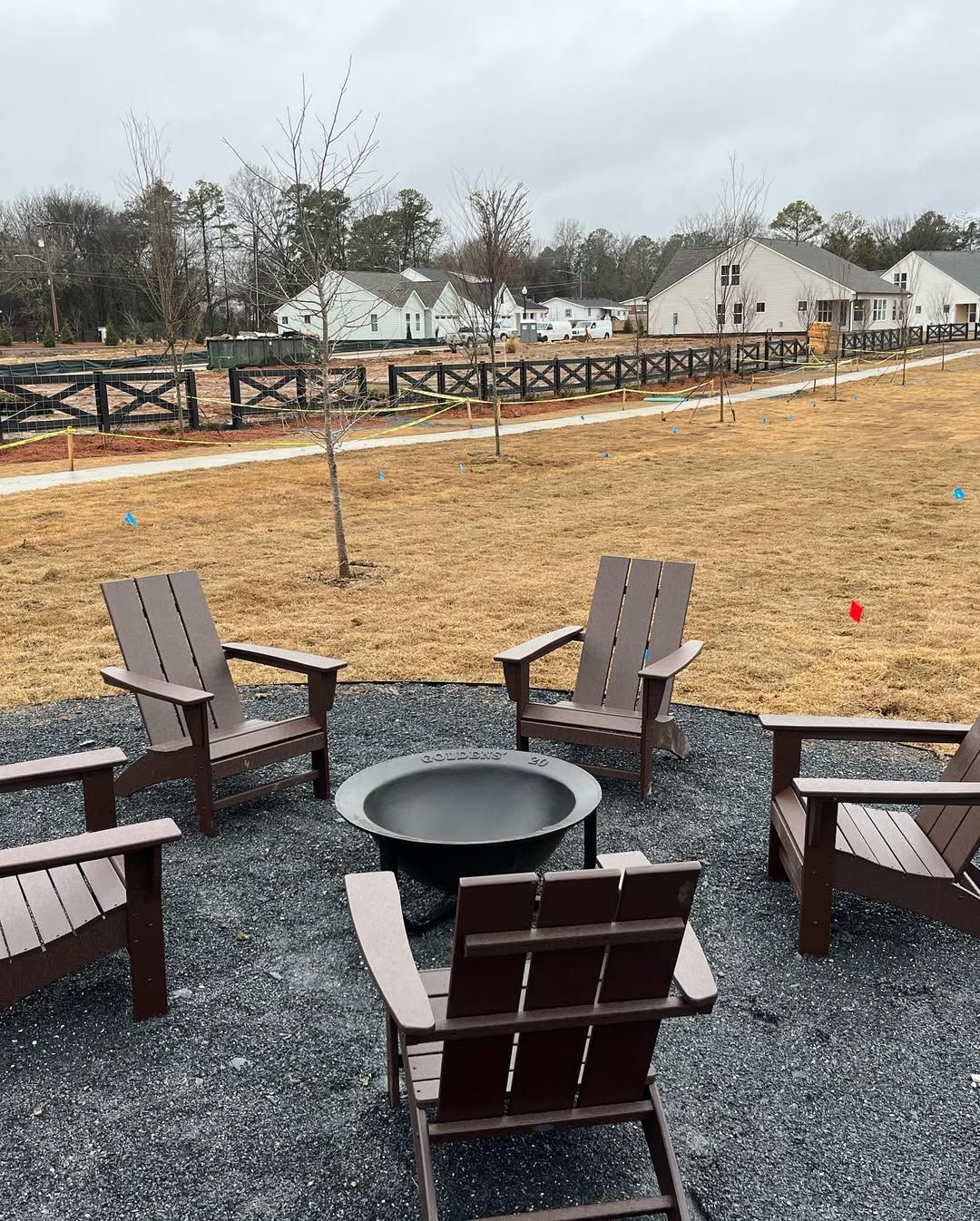 Four Adirondack chairs around a fire pit on gravel, near a path and fence, with houses in the background.