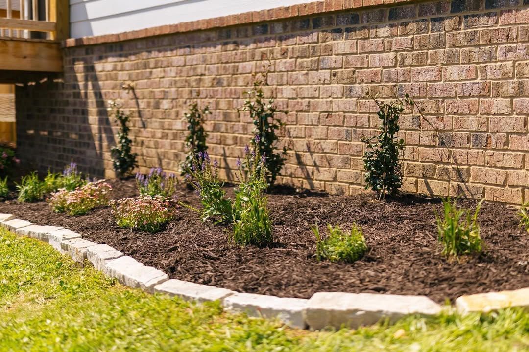 A brick house foundation with a landscaped flower bed; brown mulch and green plants.