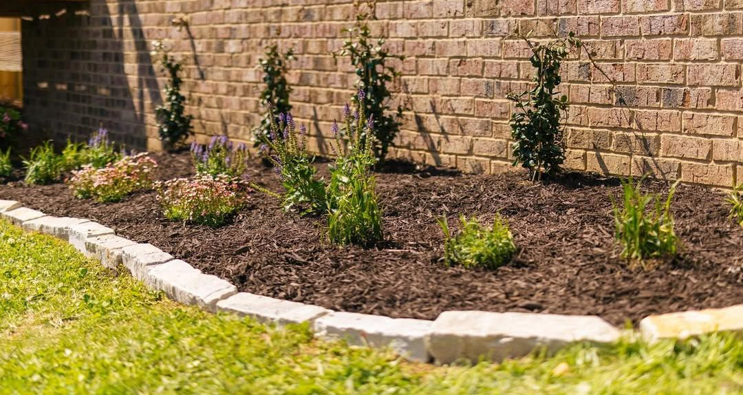 Brick wall with a flower bed, edged with stone blocks and filled with mulch and plants.