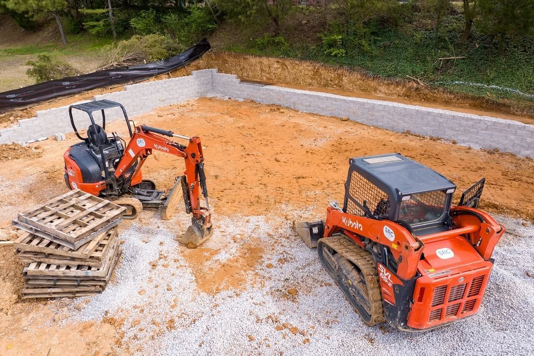 Construction site: Orange excavator and skid steer on gravel, near a foundation.