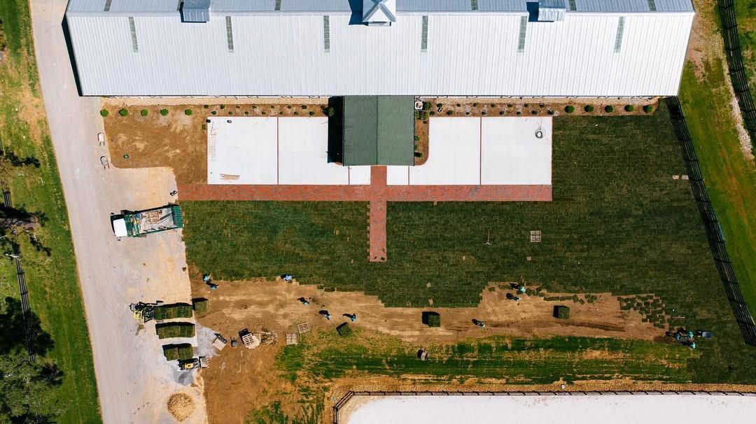 Aerial view of a building with a white roof, concrete pads, and a red brick pathway surrounded by green grass.