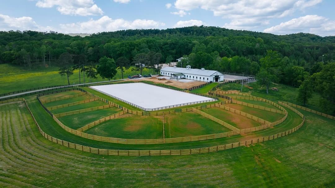 Aerial view of a white rectangular arena with tiered wooden fences in a green field, a white building in the background.