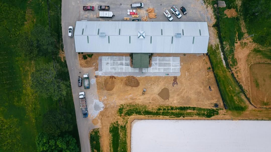 Aerial view of a long, metal-roofed building and vehicles on surrounding dirt. Green field and water in the foreground.