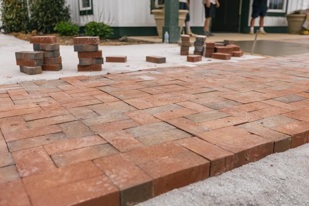 Bricks being laid in a herringbone pattern on a sandy surface, with people and a building in the background.
