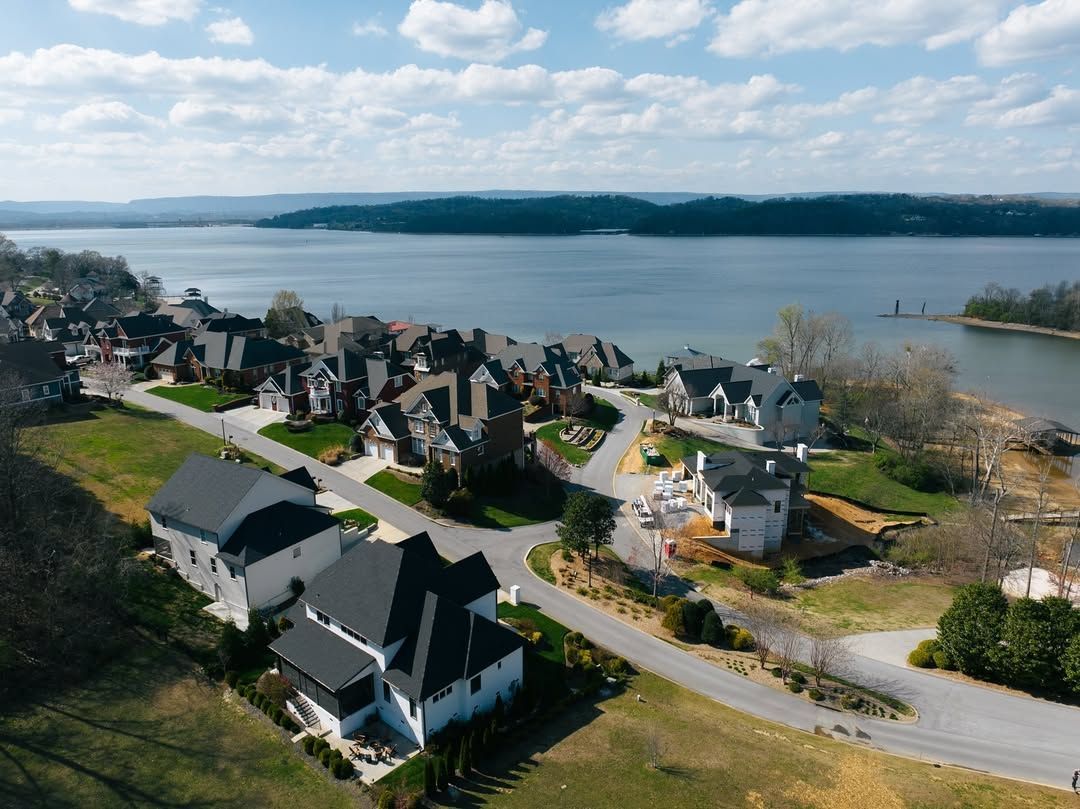 Aerial view of lakeside homes with black roofs and green lawns under a cloudy sky.