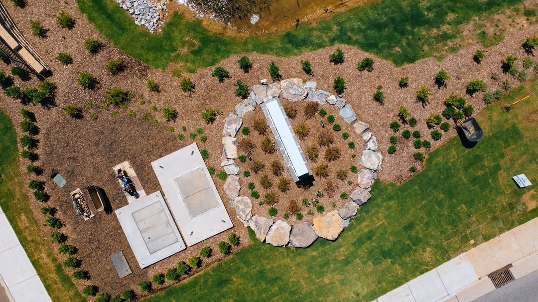 Overhead view: a sun dial surrounded by stone and plants, adjacent to concrete pads and landscaping.