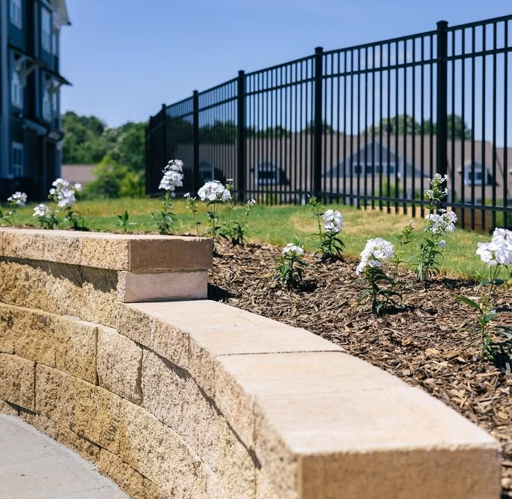 Stone retaining wall with flowers, grass, and a black fence under a blue sky.