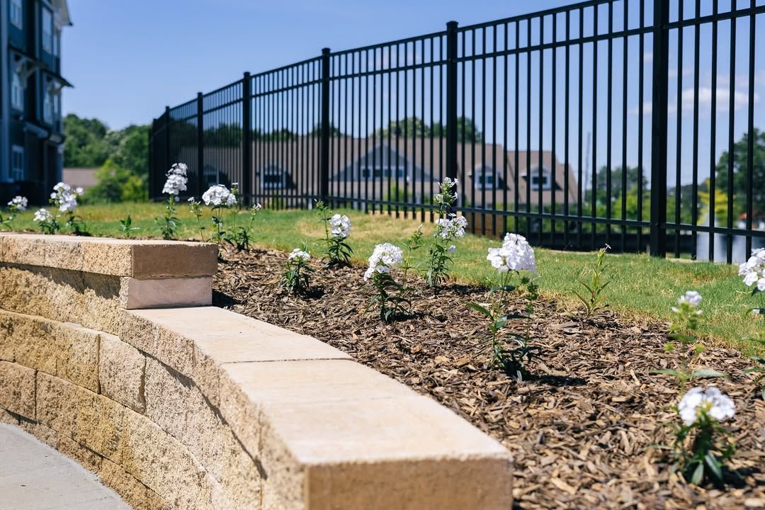 Stone retaining wall with white flowers, black metal fence, and green grass.