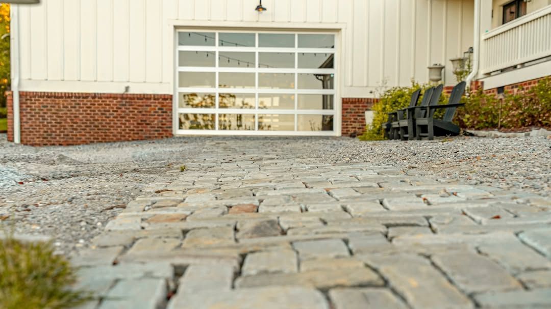 Cobblestone path leads to a white building with a glass garage door; black chairs sit nearby.