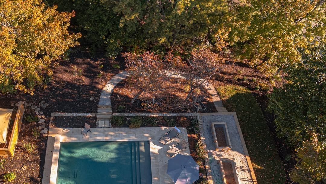 Overhead view of a swimming pool, stone patio, and pathway through a wooded yard.