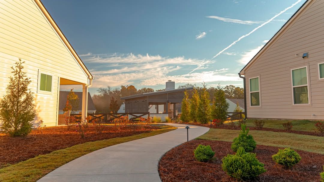 A curved pathway leads through a yard, flanked by buildings and landscaping, under a blue sky.