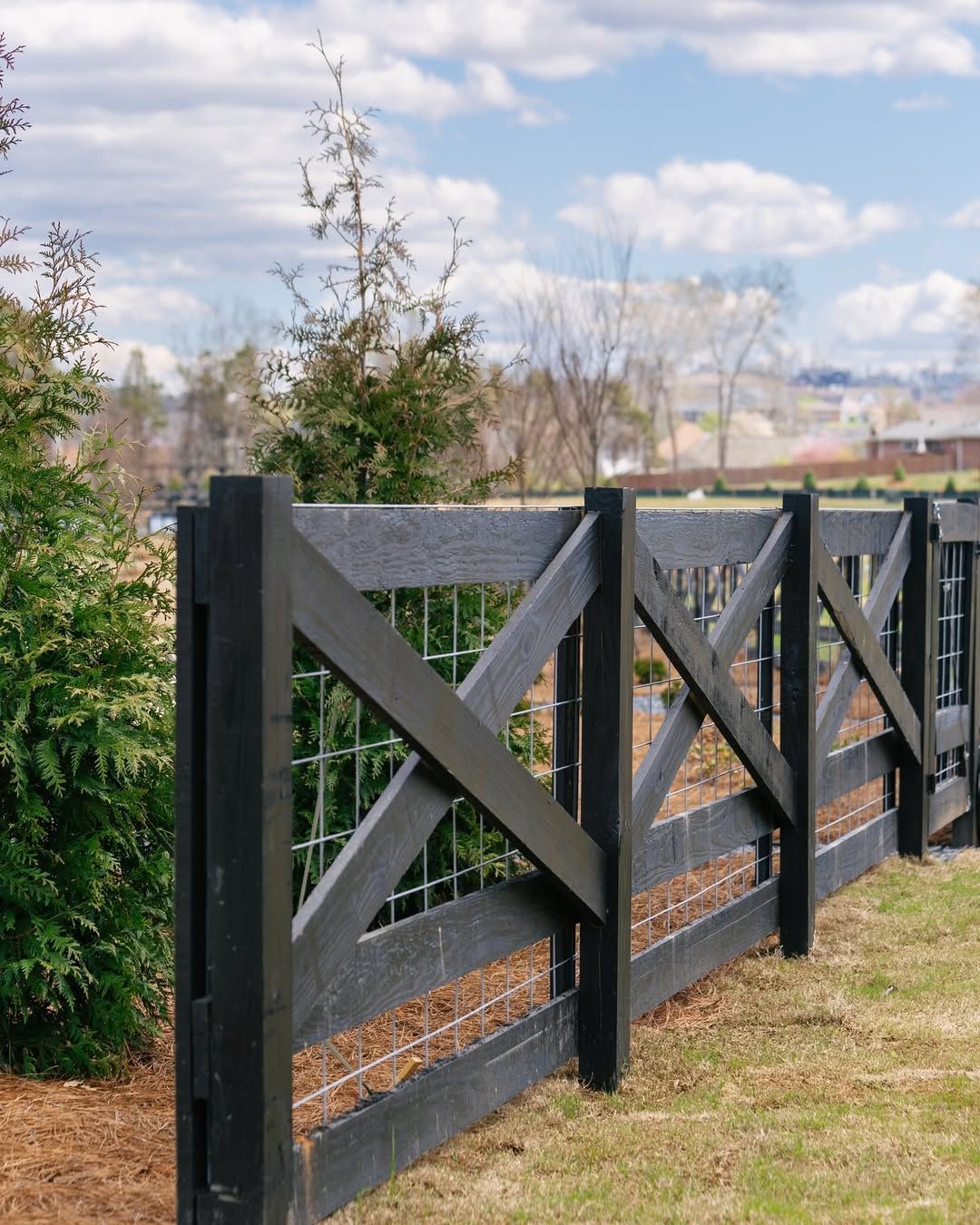 Black wooden fence with wire mesh against a grassy field and cloudy sky.