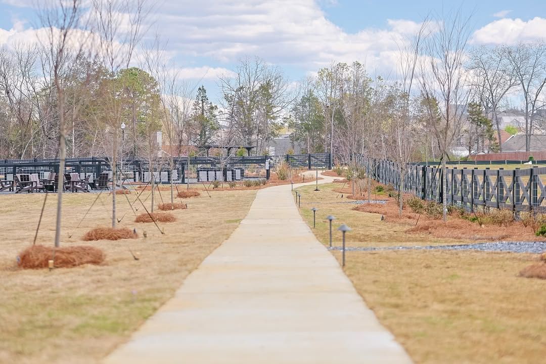 Concrete path through a park, flanked by metal fences, bare trees, and dry grass under a cloudy sky.