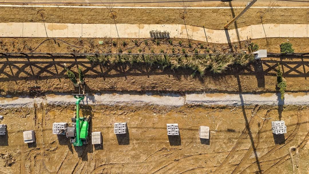 Overhead view: Construction site with a small green excavator and concrete blocks in brown dirt.