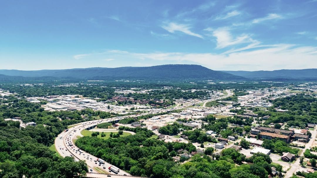 Aerial view of a city with a highway, surrounded by green trees and a mountain under a blue sky.