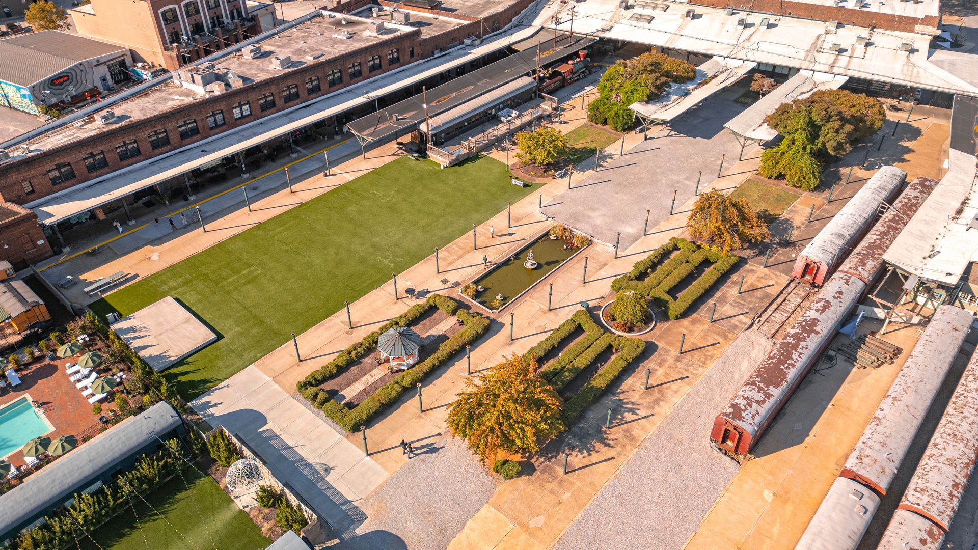 Aerial view of a plaza with grass, gardens, train cars, and brick buildings.