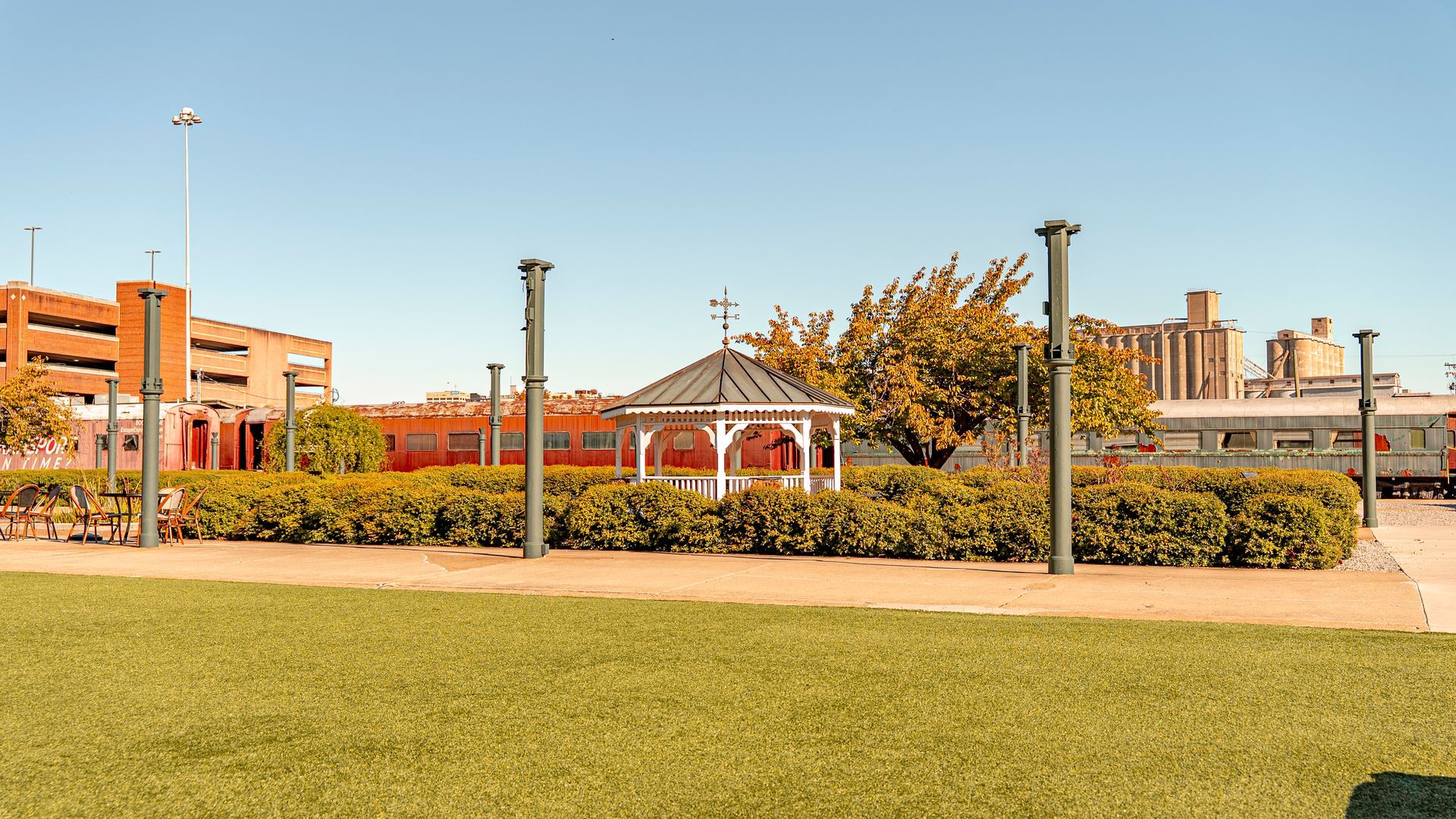 Park with a white gazebo, green bushes, and lawn. Buildings in background under a blue sky.