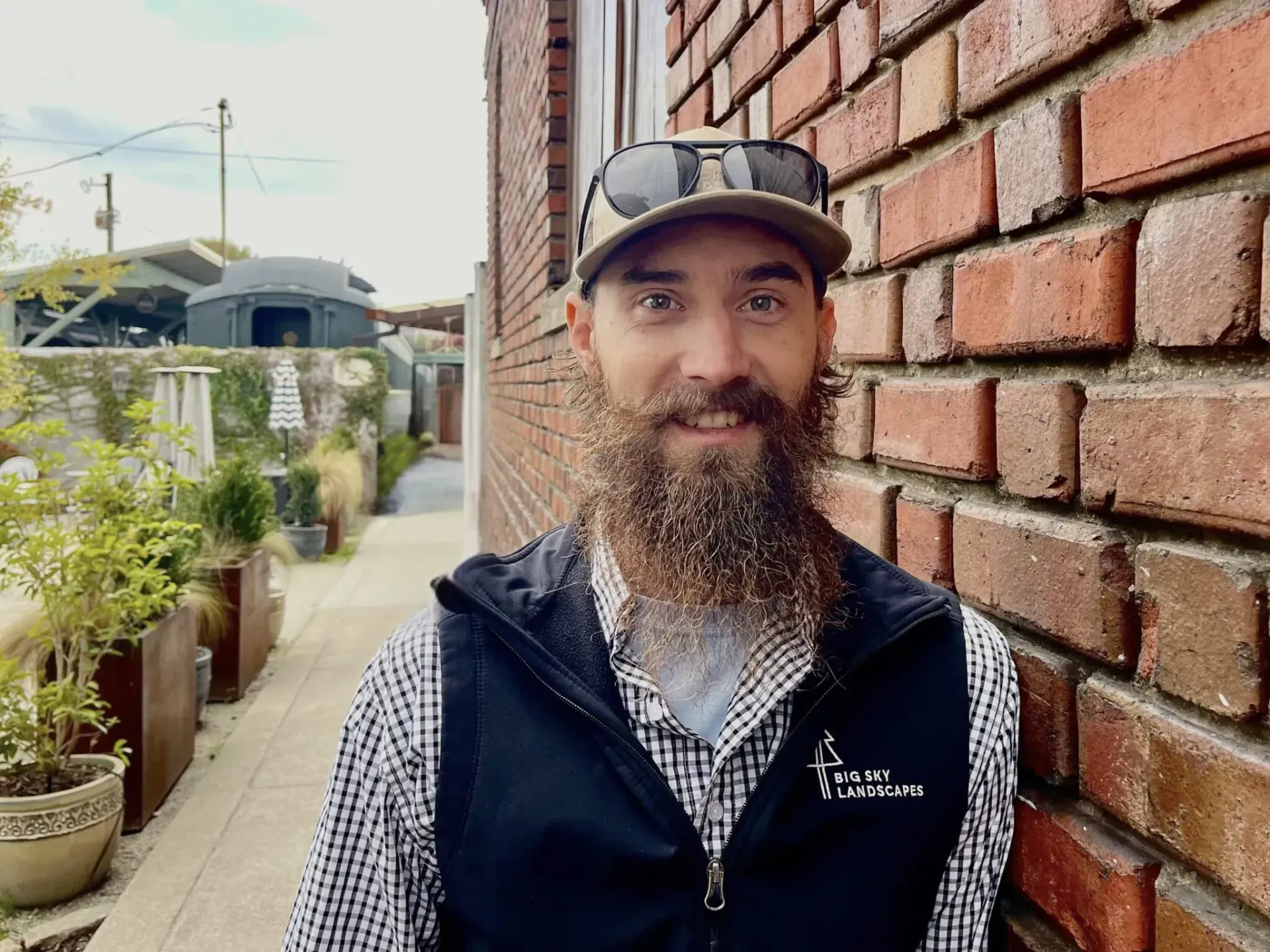 Man standing beside a brick wall on a sidewalk, wearing a cap and jacket, smiling at the camera.