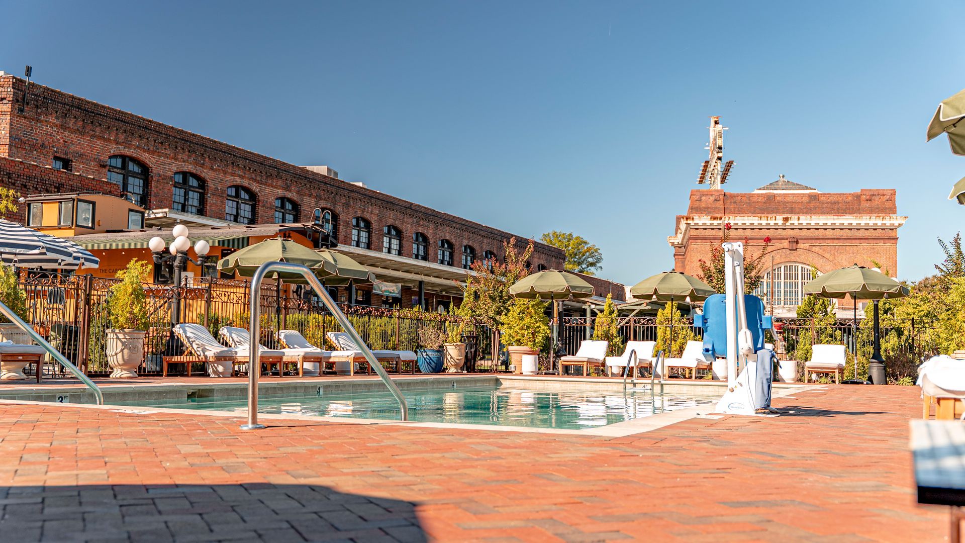 Pool with lounge chairs and umbrellas, brick buildings, sunny outdoor setting.