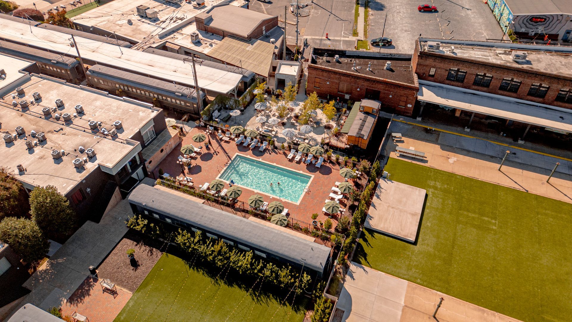 Aerial view of a rectangular pool surrounded by lounge chairs, greenery, and buildings.