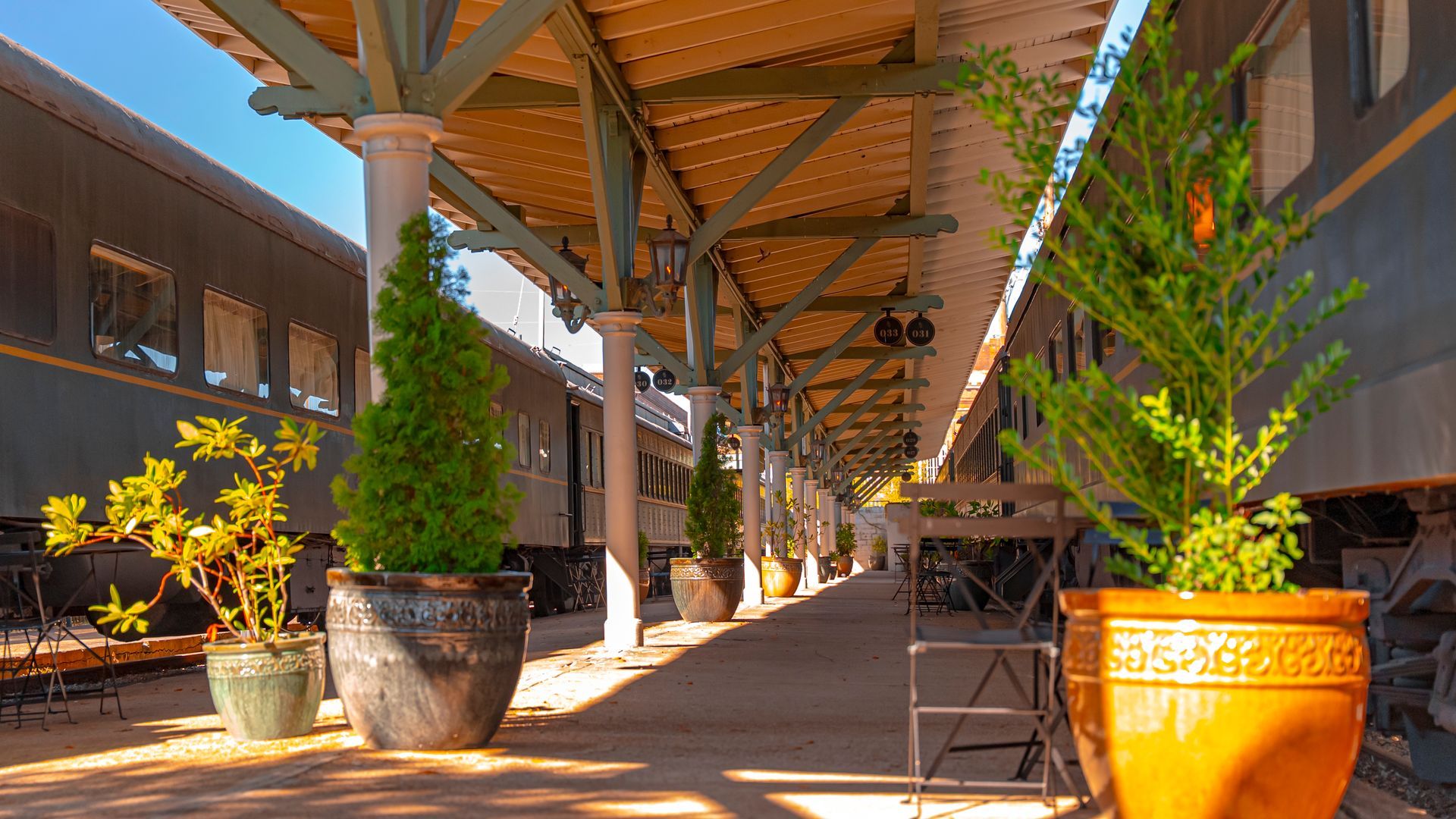Train station platform with potted plants and shade from a wooden canopy, train cars on either side.