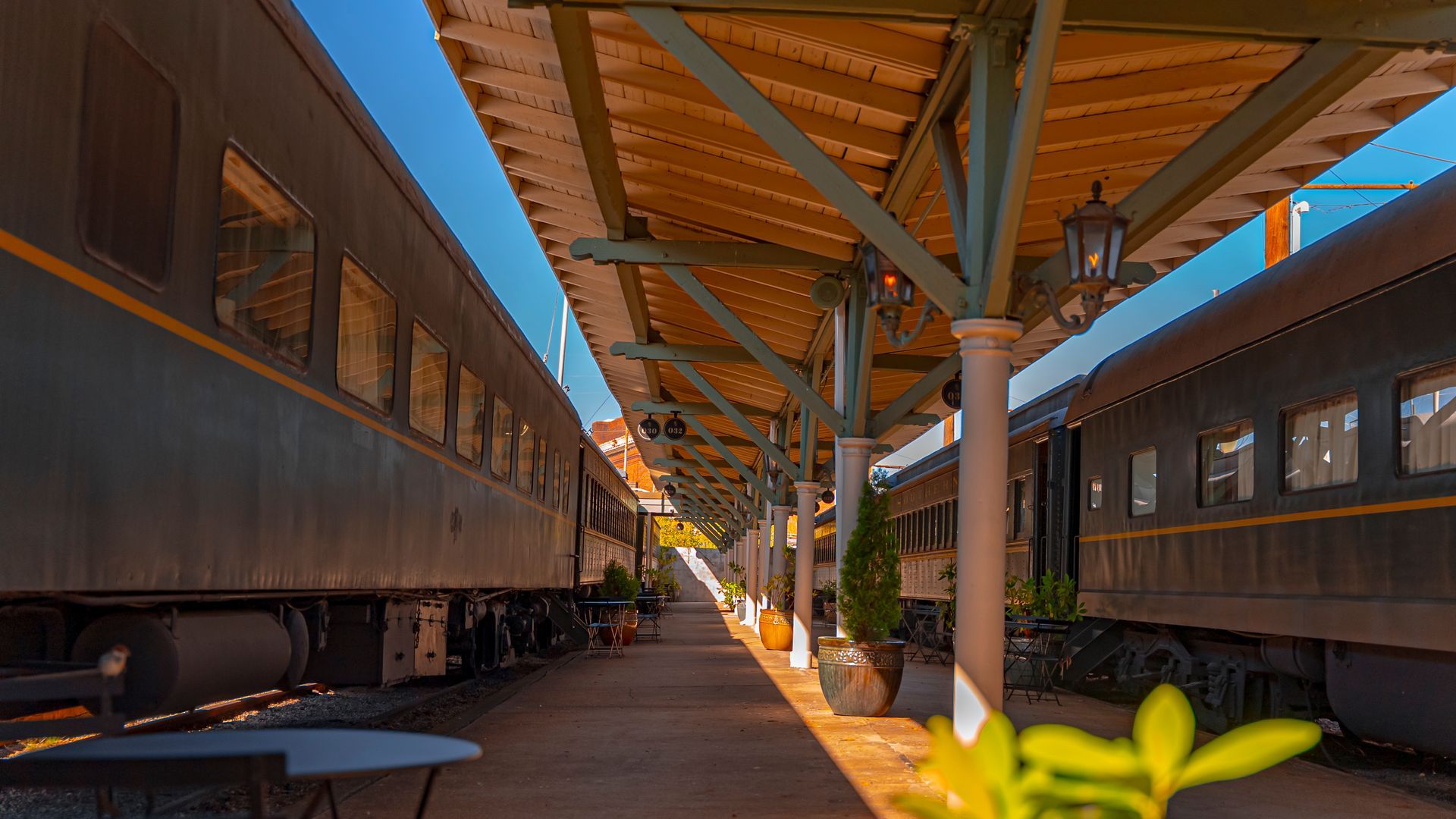 Two vintage train cars at a station with a covered platform. Sunlight casts shadows on the platform and train.