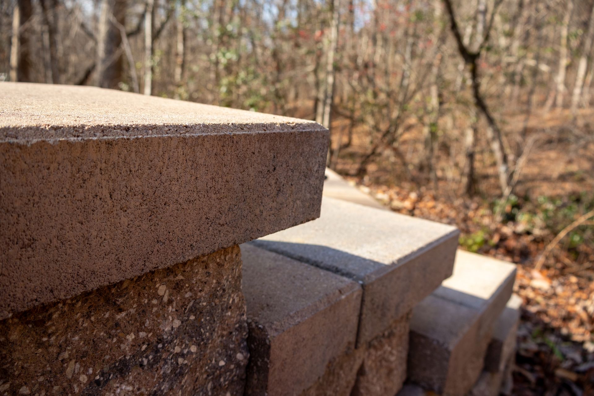 Close-up of a stacked retaining wall made of light brown concrete blocks outdoors, with blurred trees in the background.