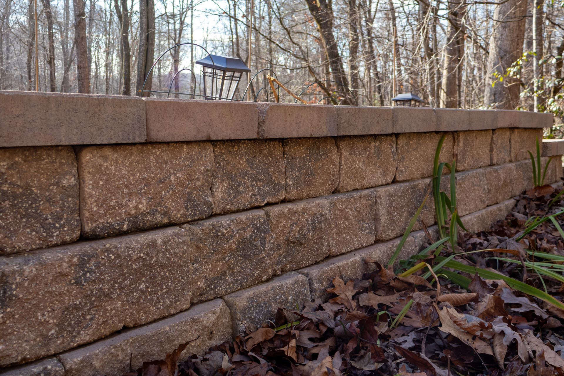 Brown brick retaining wall in a wooded area with solar lights on top.