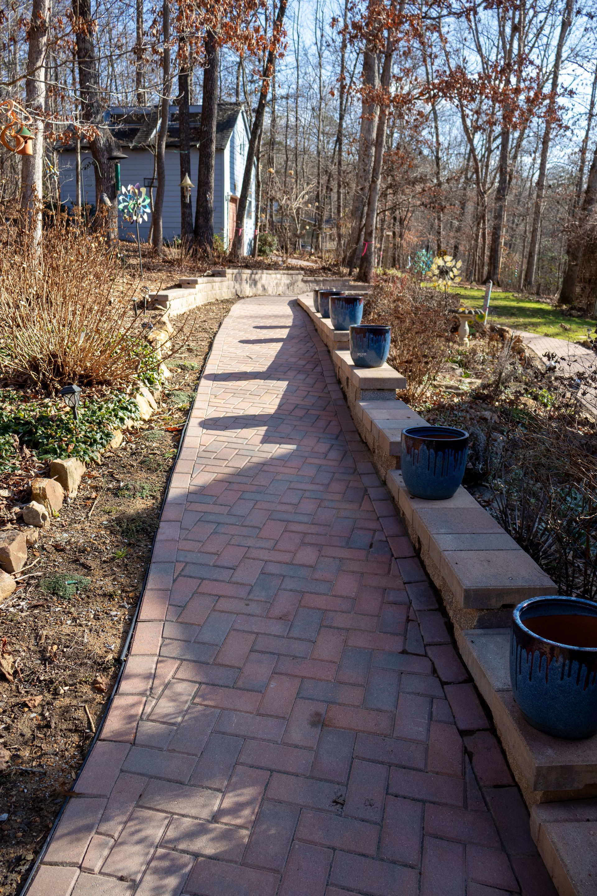 Brick pathway with blue ceramic pots on a retaining wall, leading to a shed in a wooded area.