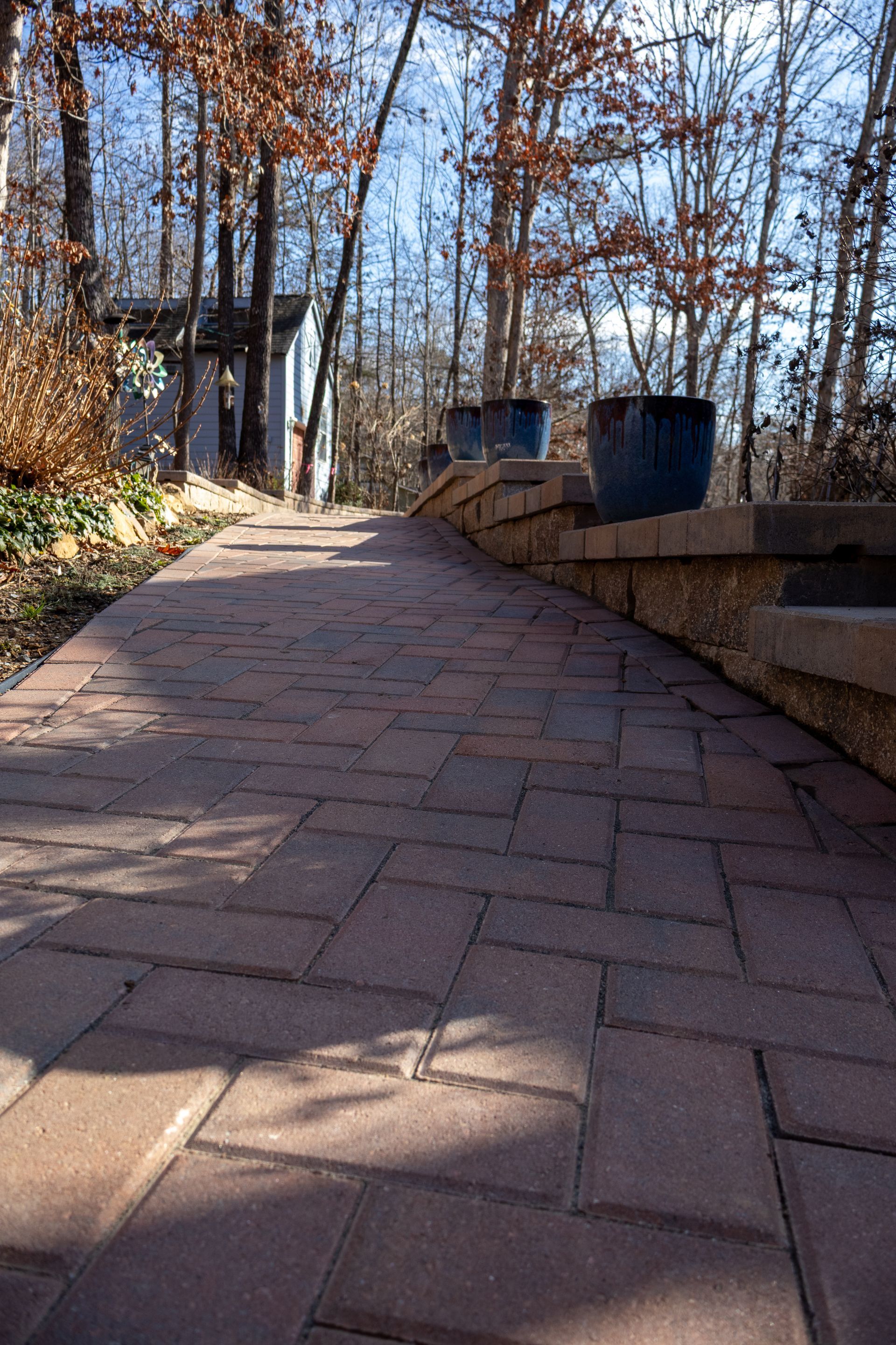 Red brick path with retaining wall, blue pots, and trees. Sunny day.