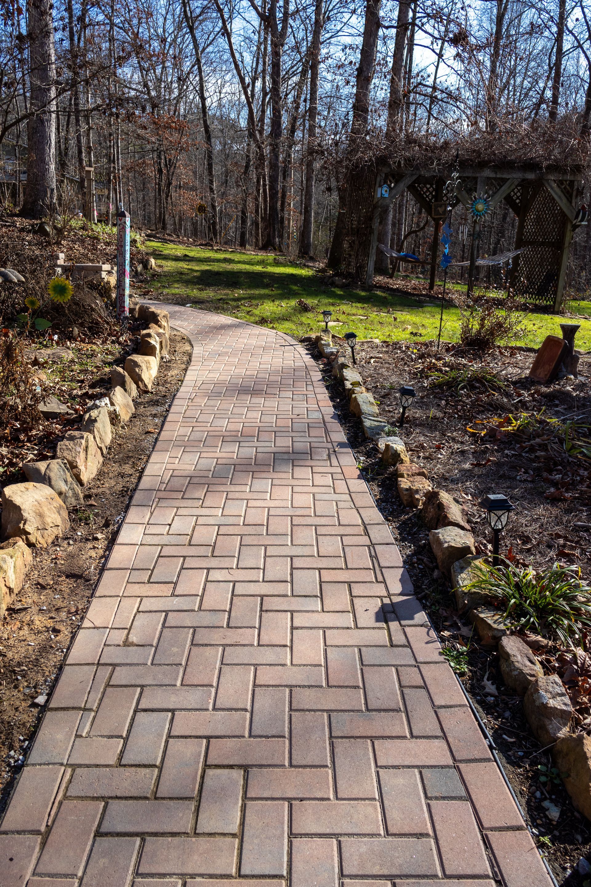 Brick pathway through a yard, bordered by rocks and plants, leading to a wooden structure.